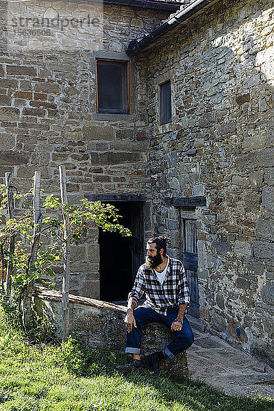 Mann mit Bart sitzt auf einer Mauer in einem rustikalen Bauernhaus