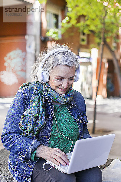 Ältere Frau mit Kopfhörern mit Laptop in der Stadt