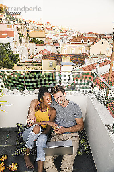 Glückliches junges Paar mit Laptop sitzt abends auf dem Dach  Lissabon  Portugal