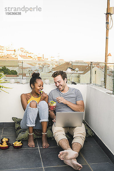 Glückliches junges Paar mit Laptop sitzt abends bei einem Imbiss auf dem Dach