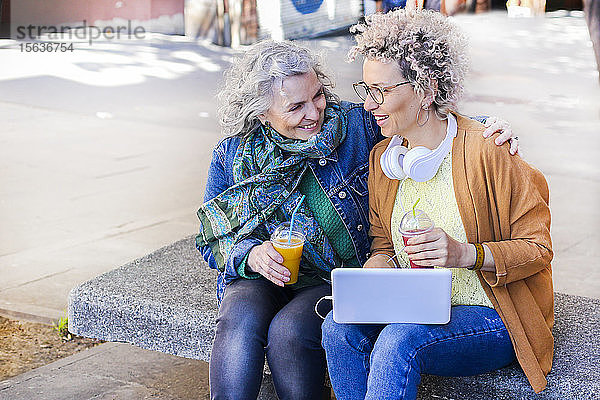 Ältere Mutter und ihre erwachsene Tochter nutzen Laptop und trinken Säfte in der Stadt