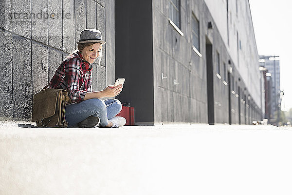 Junge Frau mit Hut sitzt an der Wand und schaut auf ein Smartphone