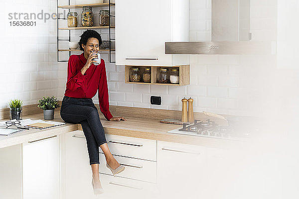 Lächelnde junge Frau sitzt zu Hause auf dem Küchentisch und trinkt aus der Tasse