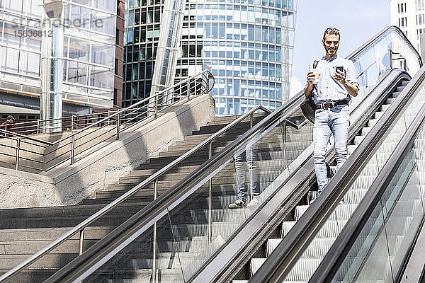 Lächelnder Geschäftsmann schaut auf das Smartphone auf der Rolltreppe in der Stadt  Berlin  Deutschland