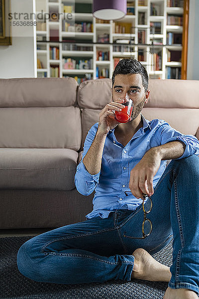 Porträt eines jungen Mannes  der zu Hause auf dem Boden sitzt und ein Glas Saft trinkt