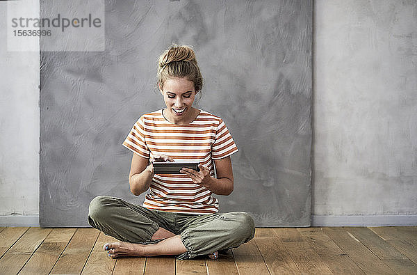 Lächelnde junge Frau sitzt auf dem Boden und benutzt ein digitales Tablett