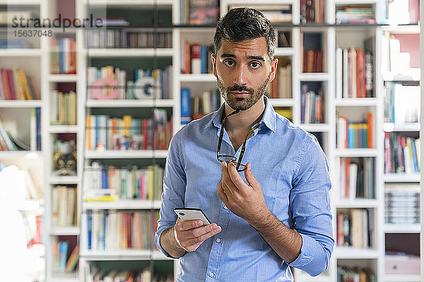 Porträt eines skeptischen jungen Mannes mit Smartphone vor einem Bücherregal stehend