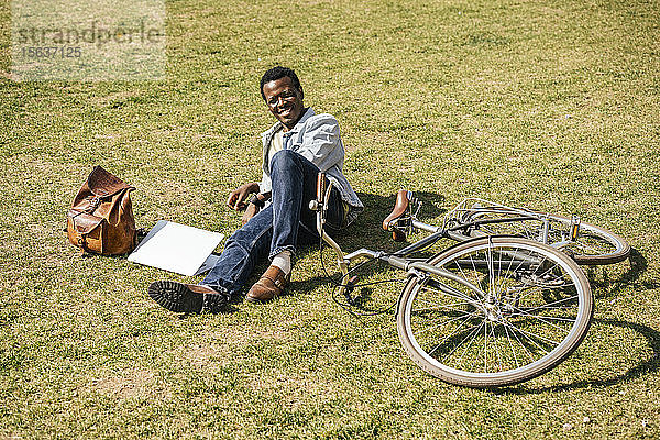 Junger Mann mit Alptop und Fahrrad  auf Gras liegend