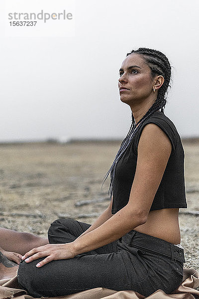 Porträt einer Frau mit schwarzen Zöpfen auf einer Decke sitzend in trostloser Landschaft  die in die Ferne schaut