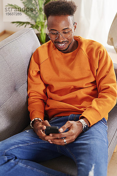 Junger lächelnder Mann sitzt auf einer Couch und benutzt ein Smartphone