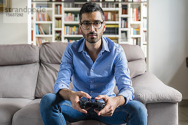 Porträt eines ernsthaften jungen Mannes  der zu Hause auf der Couch sitzt und Videospiele spielt