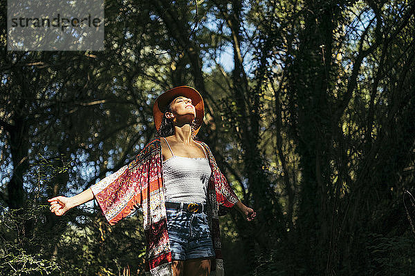 Junge Frau mit braunem Hut  farbigem Hemd und weißem Oberteil mit geschlossenen Augen und hochgekrempelten Armen  die die Sonne in einem Wald spüren