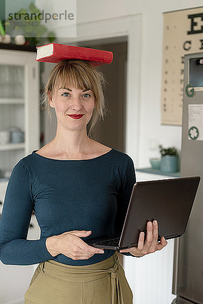 Porträt einer lächelnden Frau  die mit Laptop in der Küche steht und ein Buch auf ihrem Kopf balanciert
