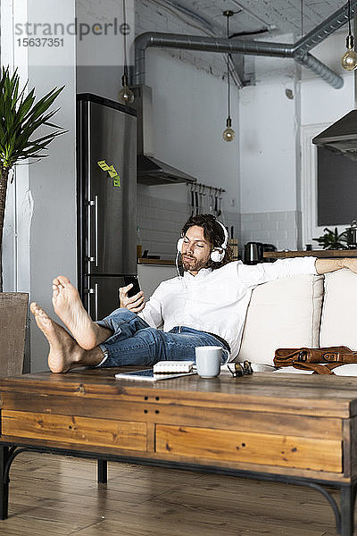 Entspannter Mann sitzt zu Hause auf der Couch mit Handy und Kopfhörern
