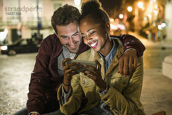 Porträt eines glücklichen jungen Paares  das bei Nacht gemeinsam auf ein Smartphone schaut  Lissabon  Portugal