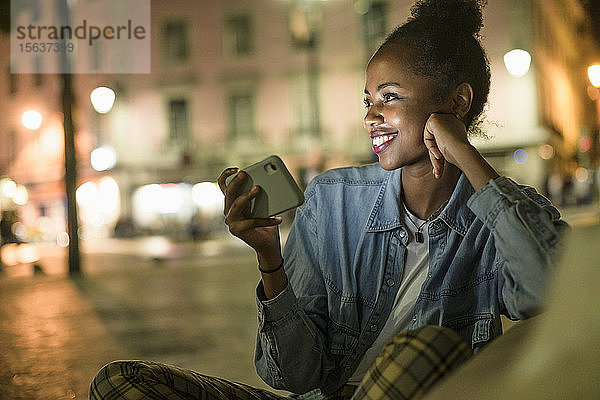 Porträt einer glücklichen jungen Frau mit Smartphone in der Stadt bei Nacht  Lissabon  Portugal