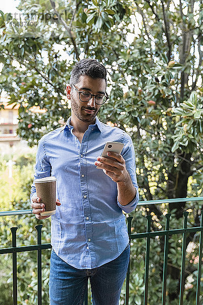 Porträt eines jungen Mannes  der mit Kaffee auf dem Balkon steht  um zum Handy zu gehen
