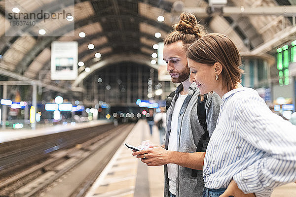 Junges Paar wartet am Bahnhof auf den Zug und benutzt ein Smartphone  Berlin  Deutschland