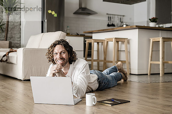 Porträt eines lächelnden Mannes  der zu Hause mit seinem Laptop auf dem Boden liegt
