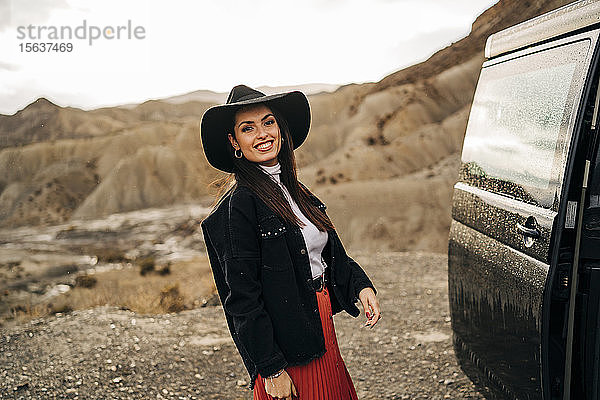 Porträt einer lächelnden jungen Frau in Wüstenlandschaft  die neben einem Wohnmobil steht  Almeria  Andalusien  Spanien