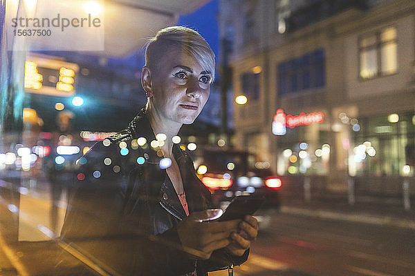 Porträt einer blonden Frau an der Straßenbahnhaltestelle am Abend mit dem Handy  Berlin  Deutschland