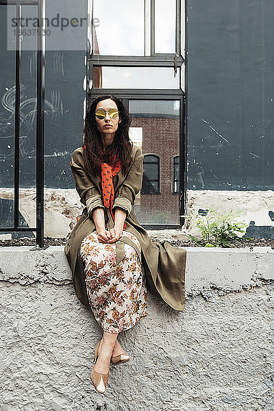 Porträt einer stilvollen Frau mit verspiegelter Sonnenbrille und rotem Schal  die an einer Wand sitzt