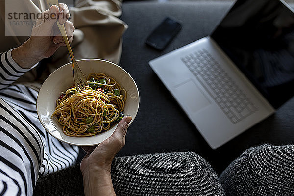 Reife Frau mit Laptop isst zu Hause auf der Couch hausgemachte Nudelgerichte