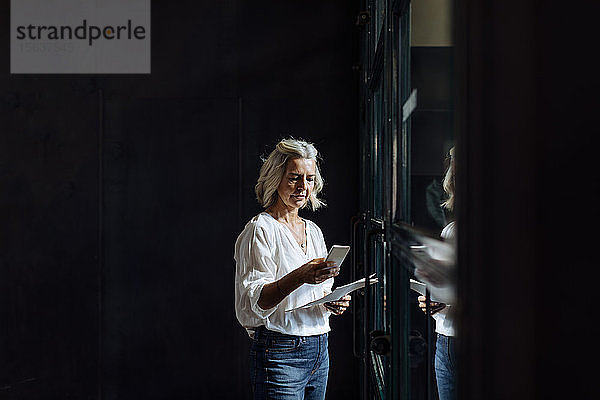 Lässig reife Geschäftsfrau mit Smartphone am Fenster im Loft-Büro