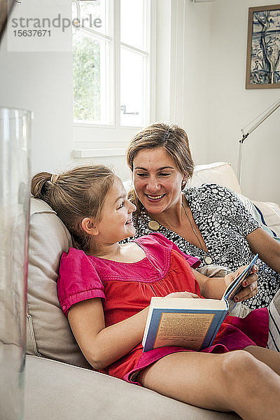 Glückliche Mutter und Tochter mit Buch auf Couch im Wohnzimmer