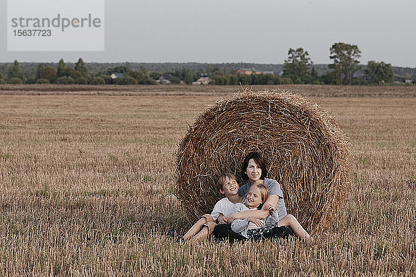 Frau mit zwei Kindern sitzt neben einem Heuhaufen auf einem Stoppelfeld
