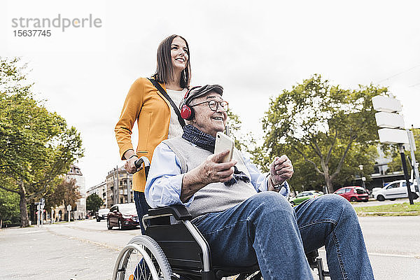 Junge Frau schiebt älteren Mann mit Kopfhörern und Smartphone im Rollstuhl