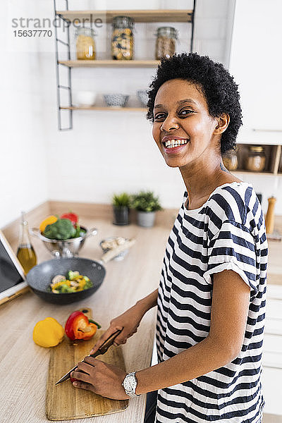 Porträt einer lächelnden jungen Frau  die zu Hause in der Küche kocht und Gemüse schneidet