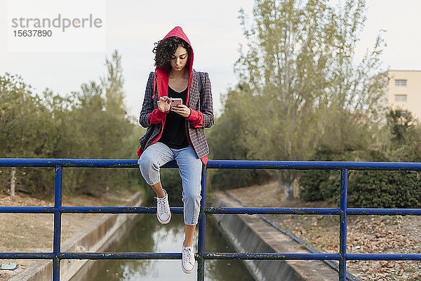 Junge Frau sitzt auf einem Geländer und benutzt ein Mobiltelefon