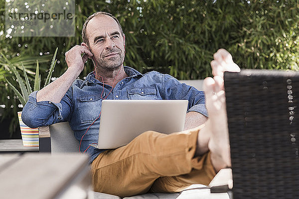 Reifer Mann sitzt mit Kopfhörern und Laptop auf der Terrasse