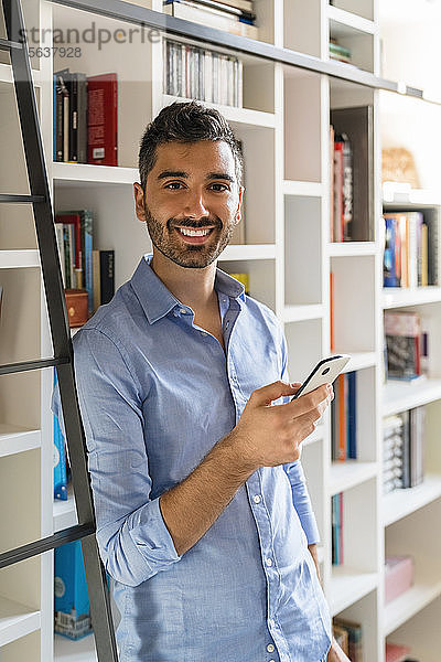 Porträt eines lächelnden jungen Mannes mit Smartphone  der zu Hause vor einem Bücherregal steht