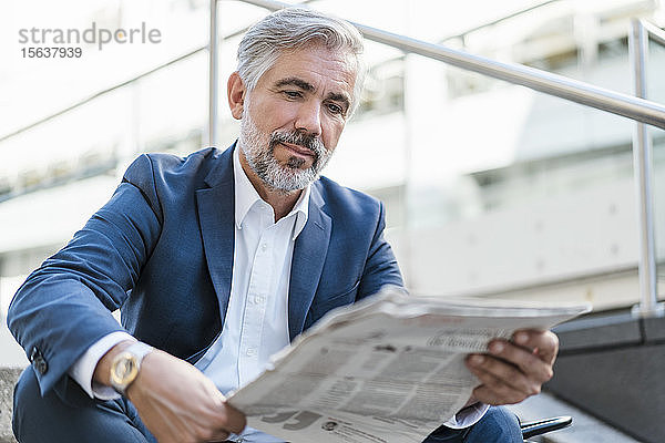 Ein reifer Geschäftsmann sitzt auf einer Treppe in der Stadt und liest Zeitung