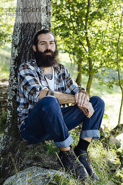 Bildnis eines Mannes mit Bart am Baumstamm im Wald sitzend