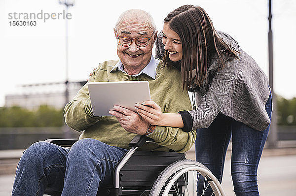 Älterer Mann sitzt im Rollstuhl und erwachsene Enkelin schauen gemeinsam auf das digitale Tablet
