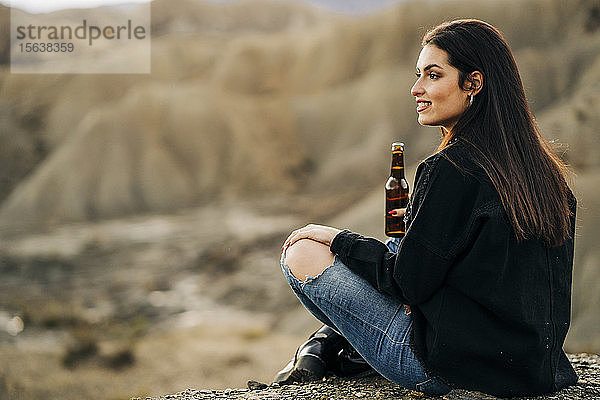 Junge Frau sitzt in Wüstenlandschaft und trinkt ein Bier  Almeria  Andalusien  Spanien