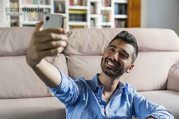 Porträt eines lächelnden jungen Mannes  der zu Hause vor der Couch sitzt und sich mit dem Handy selbstständig macht