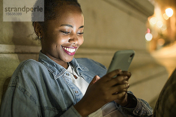 Porträt einer glücklichen jungen Frau  die ein Smartphone in der Stadt bei Nacht benutzt  Lissabon  Portugal