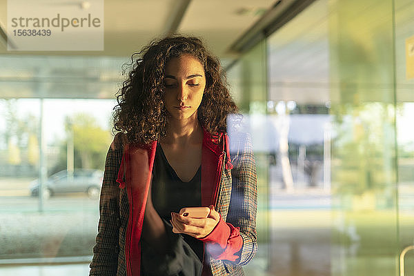 Porträt einer jungen Frau hinter Glasscheibe mit Blick auf Handy