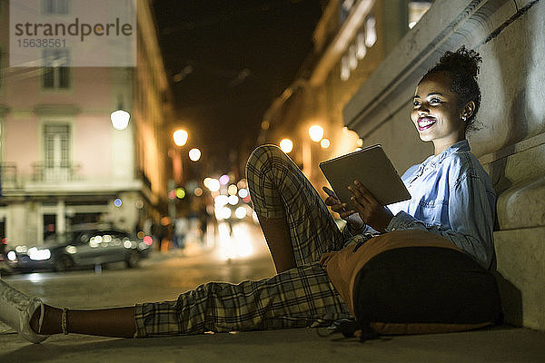 Porträt einer glücklichen jungen Frau mit Rucksack und digitalem Tablet in der Stadt bei Nacht  Lissabon  Portugal