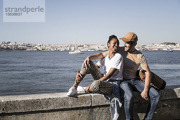 Glückliches junges Paar sitzt auf einer Mauer am Wasser  Lissabon  Portugal