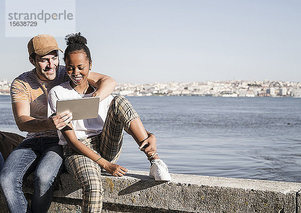 Glückliches junges Paar sitzt mit einer Tablette auf einer Mauer am Wasser  Lissabon  Portugal