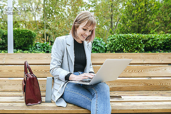 Lächelnde Frau sitzt auf Parkbank mit Laptop