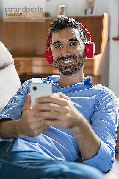 Porträt eines lächelnden jungen Mannes  der zu Hause auf der Couch liegt und ein Smartphone und drahtlose Kopfhörer benutzt