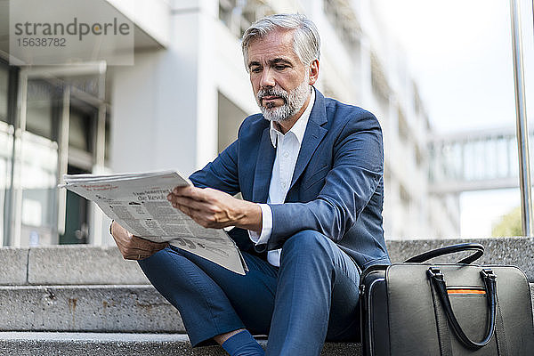 Ein reifer Geschäftsmann sitzt auf einer Treppe in der Stadt und liest Zeitung