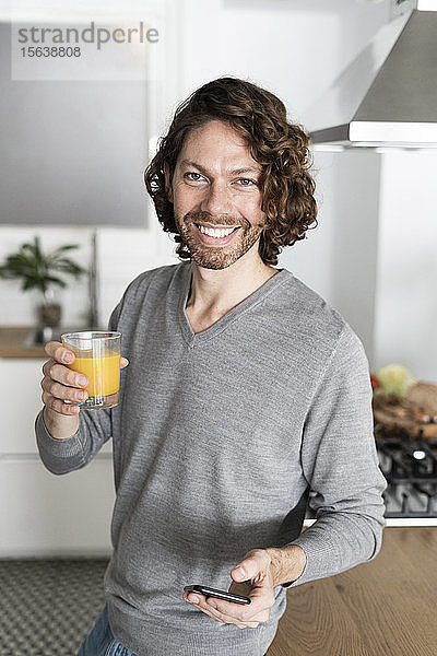 Porträt eines lächelnden Mannes mit Glas Orangensaft und Handy in der heimischen Küche