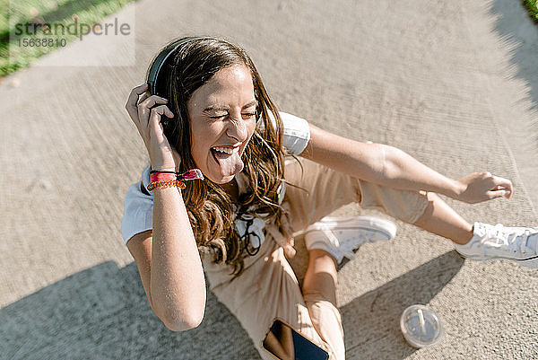 Junge Frau sitzt im Park  benutzt ein Smartphone und hört Musik mit Kopfhörern und streckt ihre Zunge heraus